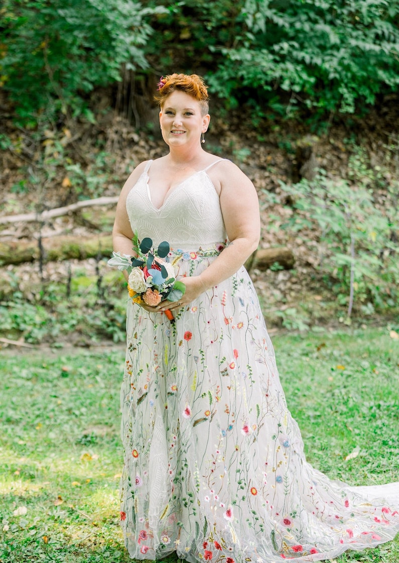 May include: A bride wearing a white wedding dress with colorful floral embroidery. She is holding a bouquet of flowers and smiling at the camera.