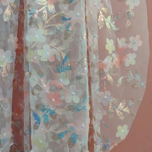 May include: Close-up of a sheer fabric adorned with delicate floral and leaf embellishments. The flowers are in shades of pink, peach, and white, with iridescent sequins. The leaves have gold and blue accents, creating a shimmering effect. The fabric is likely used for apparel or decorative purposes.