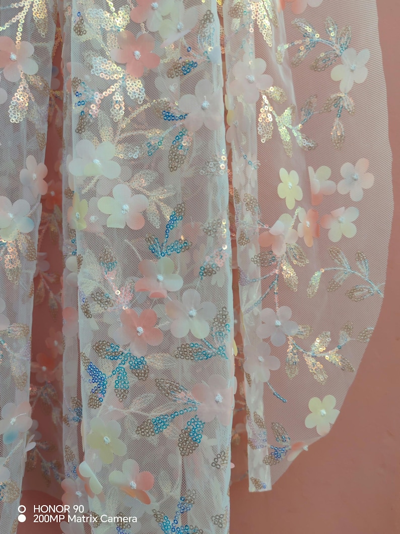 May include: Close-up of a sheer fabric adorned with delicate floral and leaf embellishments. The flowers are in shades of pink, peach, and white, with iridescent sequins. The leaves have gold and blue accents, creating a shimmering effect. The fabric is likely used for apparel or decorative purposes.