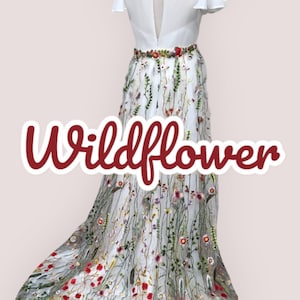 May include: A white dress with a floral embroidered skirt. The skirt features a variety of colorful flowers and greenery. The dress has a keyhole back and short sleeves. The text "Wildflower" is written in red on the skirt.