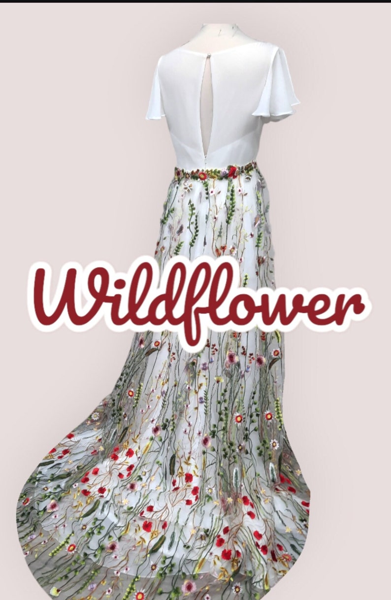 May include: A white dress with a floral embroidered skirt. The skirt features a variety of colorful flowers and greenery. The dress has a keyhole back and short sleeves. The text "Wildflower" is written in red on the skirt.