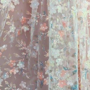 May include: Delicate floral fabric with a sheer, layered design. The fabric features white, peach, and blue flower appliqu&eacute;s, along with silver sequin accents. The background is a soft pink, creating a romantic aesthetic.