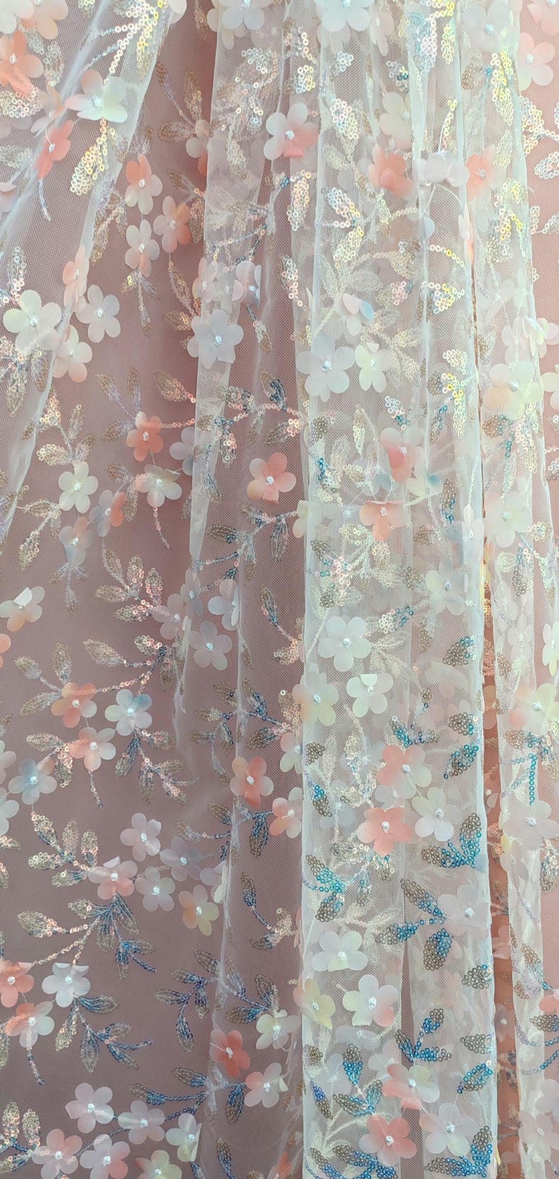 May include: Delicate floral fabric with a sheer, layered design. The fabric features white, peach, and blue flower appliqu&eacute;s, along with silver sequin accents. The background is a soft pink, creating a romantic aesthetic.