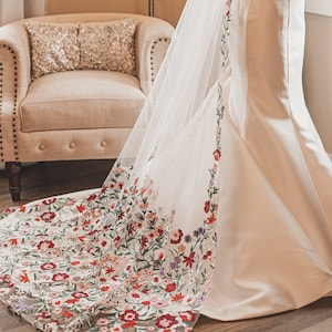 Può includere: Un abito da sposa bianco con un orlo ricamato con fiori. L'abito ha un corpetto aderente e una gonna fluida.