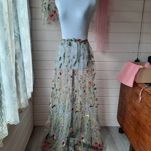 May include: A sheer tulle skirt with embroidered floral designs in shades of red, green, yellow, and blue. The skirt is displayed on a white mannequin.