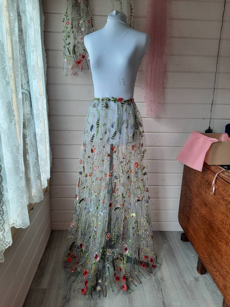 May include: A sheer tulle skirt with embroidered floral designs in shades of red, green, yellow, and blue. The skirt is displayed on a white mannequin.