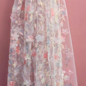 May include: Delicate white fabric with 3D floral embellishments in shades of pink, peach, and white. The floral design includes sequins and is set against a pink backdrop. This fabric is suitable for bridal wear and special occasion garments.