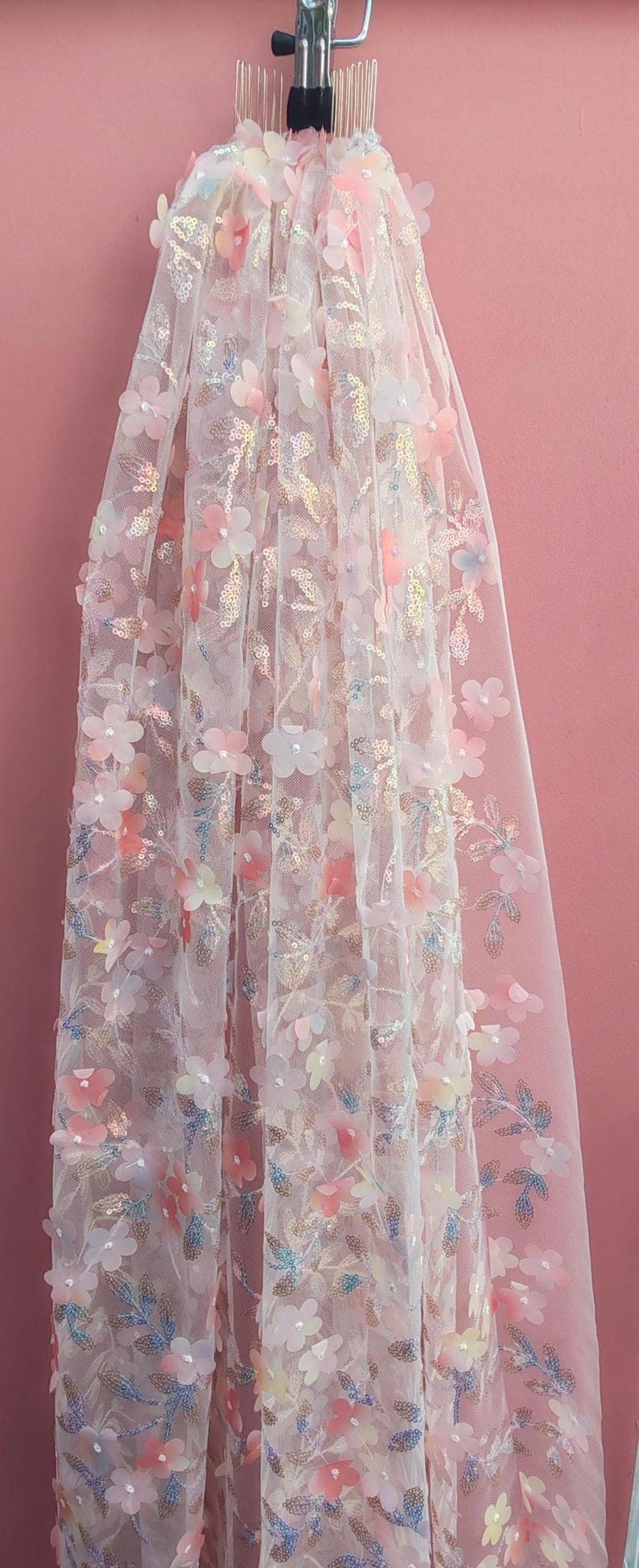 May include: Delicate white fabric with 3D floral embellishments in shades of pink, peach, and white. The floral design includes sequins and is set against a pink backdrop. This fabric is suitable for bridal wear and special occasion garments.