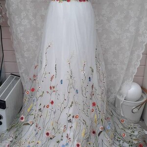 May include: A white tulle wedding dress with a low back and a floral embroidery design. The dress has a fitted bodice and a flowing skirt with a delicate floral pattern.