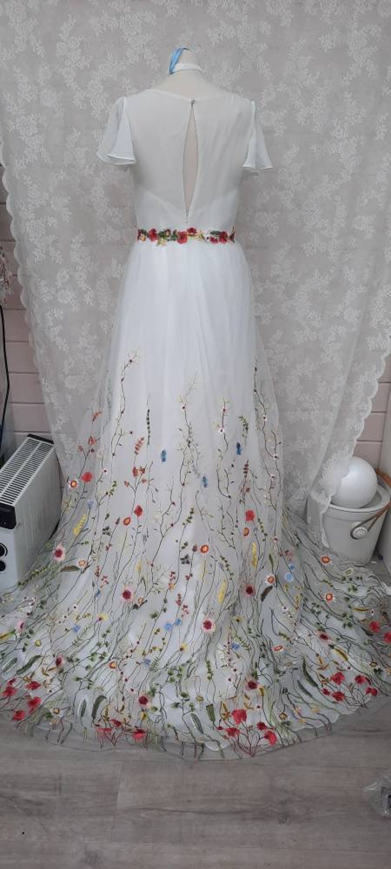 May include: A white tulle wedding dress with a low back and a floral embroidery design. The dress has a fitted bodice and a flowing skirt with a delicate floral pattern.