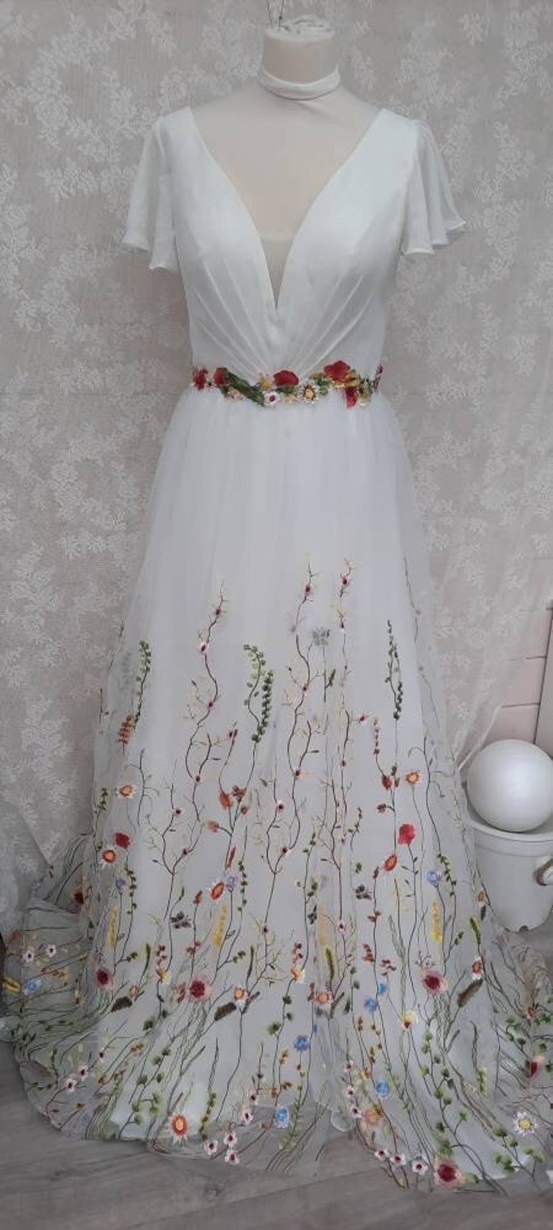 May include: A white wedding dress with a V-neckline and short sleeves. The dress is adorned with a floral embroidery pattern in shades of red, pink, yellow, and blue.