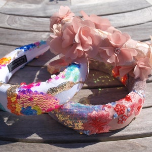 Könnte beinhalten: Drei Stirnbänder in verschiedenen Ausführungen. Ein Stirnband ist rosa mit rosa Stoffblumen, eines ist weiß mit bunten Pailletten und eines ist rosa mit roten gestickten Blumen.