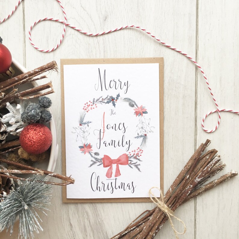May include: A Christmas card featuring a watercolour wreath design and the words "Merry The Jones Family Christmas." The card is surrounded by festive decorations, including red and white twine, a red bauble, and pine cones.