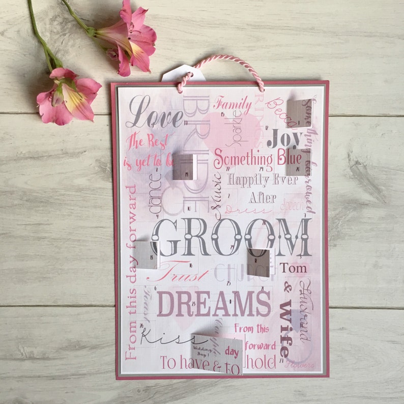 May include: A pink and white wedding sign with the word "GROOM" in the center. The sign is decorated with various wedding-related words and phrases, including "Love", "Family", "Something Blue", "Happily Ever After", "Dreams", "Kiss", "To Have & To Hold", and "Wedding Day".