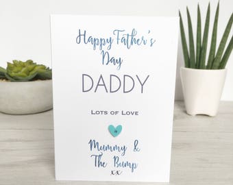 May include: A white card with blue text that reads "Happy Father's Day Daddy Lots of Love Mummy & The Bump xx". A light blue heart with a white button is attached to the card.
