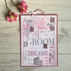 May include: A pink and white wedding sign with the word "GROOM" in large letters. The sign is decorated with various wedding-related words and phrases, including "Love", "Family", "Something Blue", "Happily Ever After", "Trust", "Dreams", "Kiss", "To Have & To Hold", and "Husband & Wife".