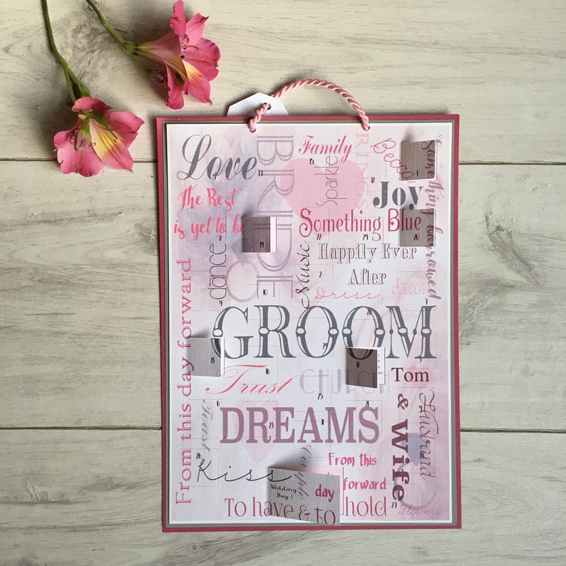 May include: A pink and white wedding sign with the word "GROOM" in large letters. The sign is decorated with various wedding-related words and phrases, including "Love", "Family", "Something Blue", "Happily Ever After", "Trust", "Dreams", "Kiss", "To Have & To Hold", and "Husband & Wife".