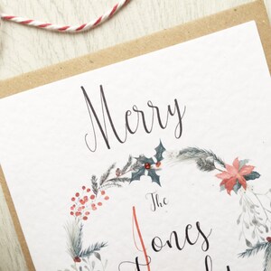 May include: A festive Christmas card with a watercolour wreath design. The card displays the words "Merry" and "The Jones" in elegant script. The wreath is decorated with red berries, holly, and other seasonal greenery, set against a white background.
