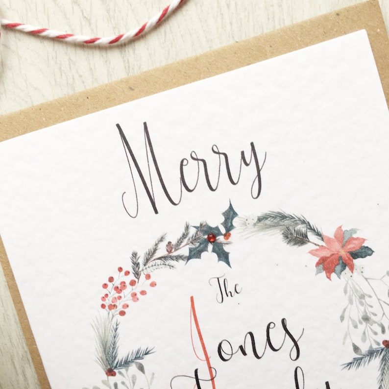 May include: A festive Christmas card with a watercolour wreath design. The card displays the words "Merry" and "The Jones" in elegant script. The wreath is decorated with red berries, holly, and other seasonal greenery, set against a white background.