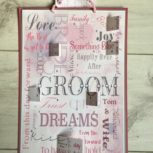 May include: A pink and white card with the word "GROOM" in large letters. The card has a variety of other words and phrases printed on it, including "Love", "Family", "Something Blue", "Happily Ever After", "Trust", "Dreams", "Kiss", "To Have & To Hold", and "Wedding Day".