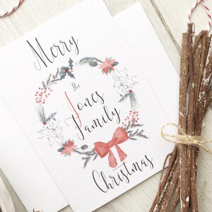 May include: A white Christmas card with a watercolour wreath design. The wreath features red berries, poinsettias, and a red bow. The card reads "Merry The Jones Family Christmas" in elegant script.