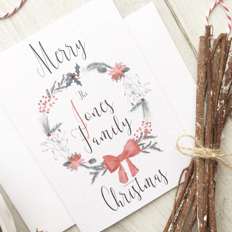 May include: A white Christmas card with a watercolour wreath design. The wreath features red berries, poinsettias, and a red bow. The card reads "Merry The Jones Family Christmas" in elegant script.