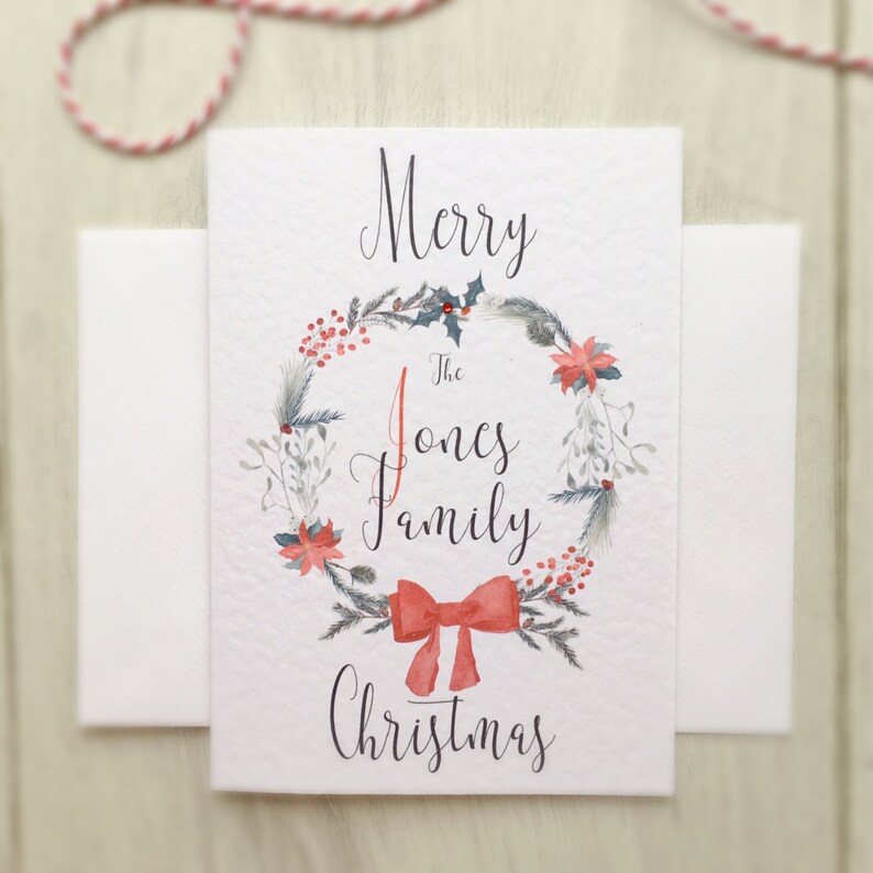 May include: A white Christmas card with a watercolour wreath design. The wreath features red berries, green leaves, and a red bow. The card reads "Merry The Jones Family Christmas" in elegant script.