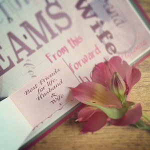 May include: A pink and white flower with green leaves sits on a wooden surface. A card with the text "Best Friends for life, Husband & Wife" is visible in the background.