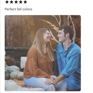 May include: A couple sitting on a hay bale in a field with fall foliage. The woman is wearing a brown sweater and blue jeans. The man is wearing a blue shirt and jeans. They are looking at each other and smiling.