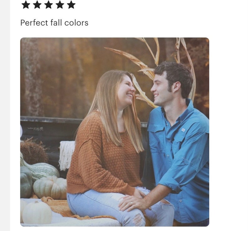May include: A couple sitting on a hay bale in a field with fall foliage. The woman is wearing a brown sweater and blue jeans. The man is wearing a blue shirt and jeans. They are looking at each other and smiling.