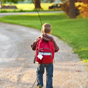 May include: A young person wearing a red backpack with a white tag walks down a gravel path. The path is lined with green grass and trees.