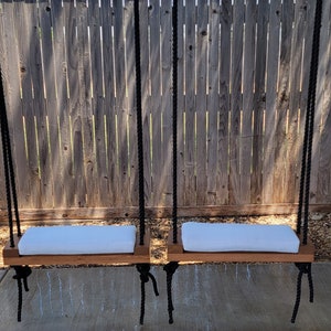 May include: Two wooden swing seats with white cushions hanging from black ropes in front of a wooden fence.