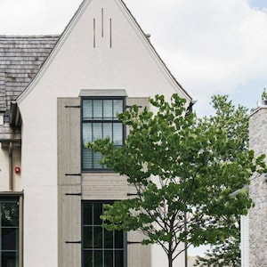 May include: A white house with a gray roof and a large window with wooden shutters. A tree is in front of the house.