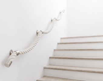 May include: A white rope handrail with silver metal hardware is mounted on a white wall. The handrail curves along the wall and over a set of white stairs with light brown treads. The design is minimalist and nautical.