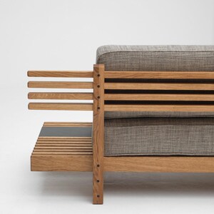 May include: A wooden sofa with a gray fabric cushion and a slatted back and armrests. The sofa has a small shelf underneath the seat.
