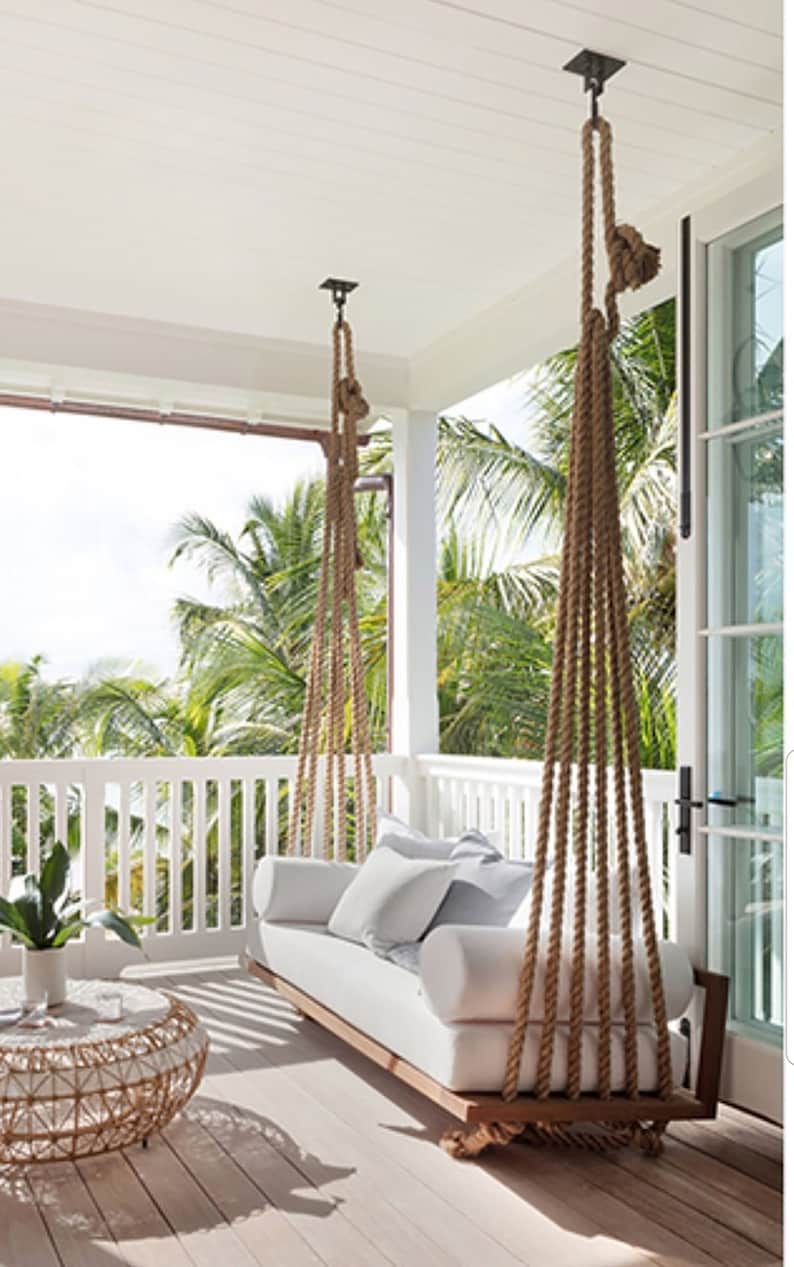 May include: A white cushioned swing bed hangs from the ceiling of a porch with a white railing. The swing bed is made of wood and has a white cushion. The porch has a wooden floor and a white railing. There are palm trees in the background.