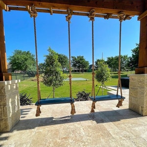 May include: Three wooden porch swings with dark blue seats hang from thick, knotted ropes attached to a wooden beam. The swings are suspended above a stone patio, with a green lawn and trees in the background under a bright blue sky.