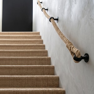 May include: A staircase with beige carpeted steps and a natural rope handrail. The handrail is supported by black metal brackets. A black square is visible on the wall at the top of the stairs.