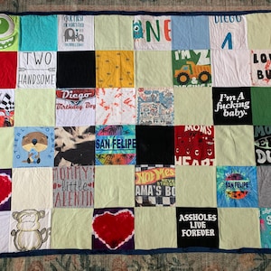 May include: A memory quilt constructed from various colorful t-shirt squares featuring diverse graphic prints, including cartoon characters, hearts, and text such as "Two Handsome," "Mom's Epic Dude," and "Assholes Live Forever," finished with a dark blue border.