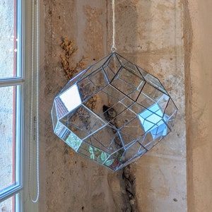 Può includere: Un terrario di vetro geometrico appeso a una corda. Il terrario è fatto di vetro trasparente e ha una forma di diamante. È appeso in una stanza con una parete bianca e una trave di legno.