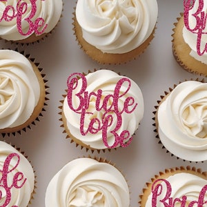 Könnte beinhalten: Eine Schachtel Cupcakes, die mit pinken Glitzer-Toppern 'Bride to Be' verziert sind. Die Cupcakes sind weiß mit einem Wirbeldesign.