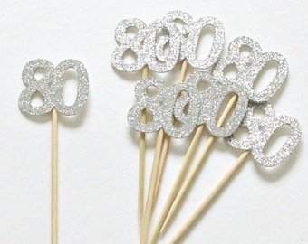 May include: Silver glitter number 80 cake toppers on wooden sticks. There are 10 toppers in total.