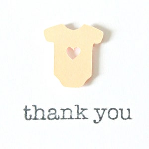 May include: A light yellow paper cutout of a baby onesie with a heart-shaped hole in the center. The text "thank you" is printed below the onesie.