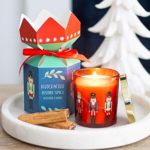 Può includere: Una candela accesa in vetro arancione con coperchio dorato, con illustrazioni di schiaccianoci, è su un vassoio bianco con una scatola decorativa. La scatola è blu e rossa, con le parole "Nutcracker Festive Spice Scented Candle". Due bastoncini di cannella sono davanti.