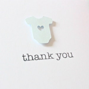 May include: A white card with a light blue paper cutout of a baby onesie with a heart cutout. The card says "thank you" in black text.