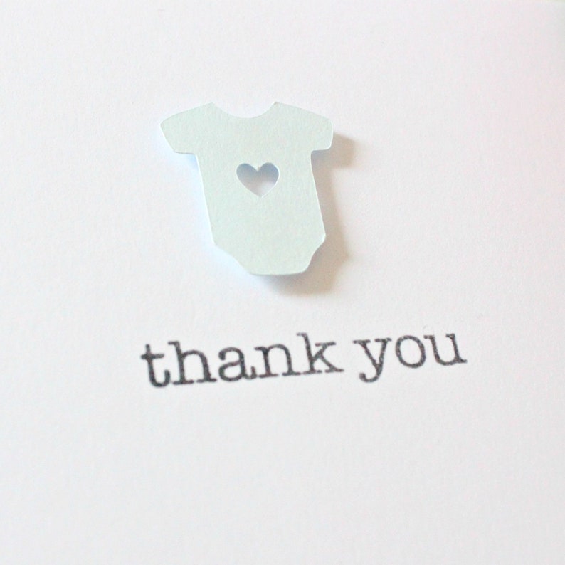 May include: A white card with a light blue paper cutout of a baby onesie with a heart cutout. The card says "thank you" in black text.