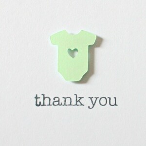 May include: A green paper cutout of a baby onesie with a heart cutout in the center. The text "thank you" is printed below the onesie.