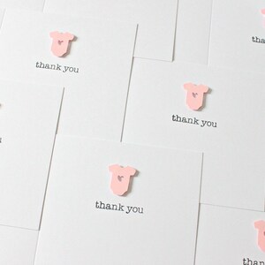 May include: A set of pink thank you cards with a small pink baby onesie with a heart on it. The cards say "thank you" in black text.