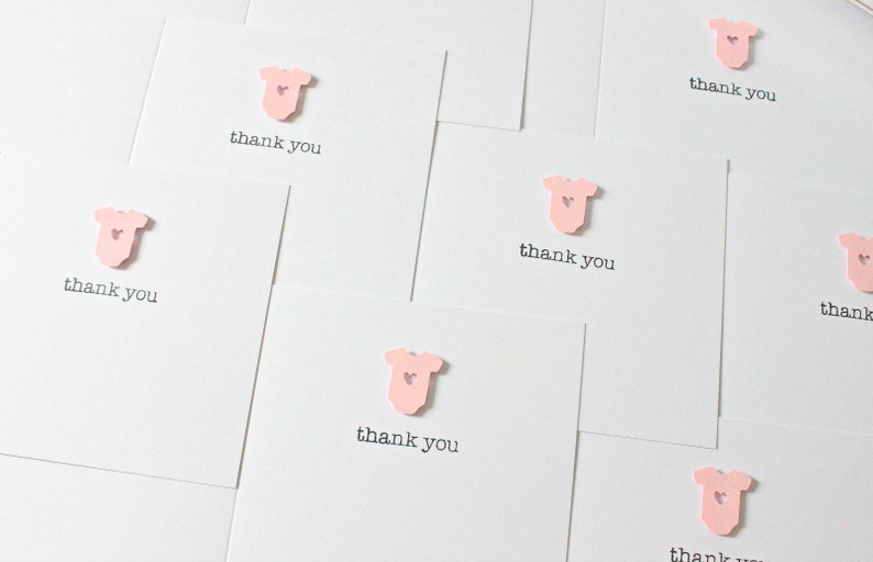 May include: A set of pink thank you cards with a small pink baby onesie with a heart on it. The cards say "thank you" in black text.