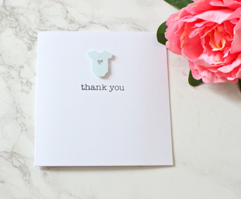May include: A white card with a light blue baby onesie cutout with a small heart in the center. The text "thank you" is printed below the onesie.